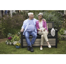 Un cuplu în vârstă stă pe o bancă de grădină cu spațiu de depozitare, înconjurat de plante.