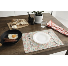 Suport de farfurie floral pe o masă din lemn închis, cu o farfurie albă, tacâmuri, o tigaie cu ou, pâine și o plantă de apartament.