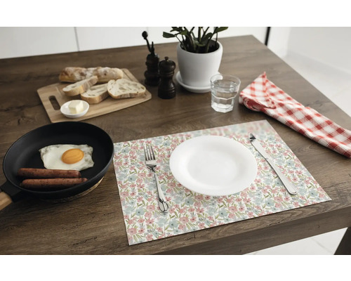 Suport de farfurie floral pe o masă din lemn închis, cu o farfurie albă, tacâmuri, o tigaie cu ou, pâine și o plantă de apartament.