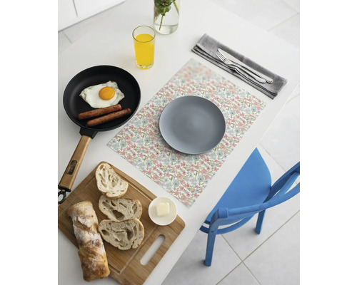 Suport de farfurie cu imprimeu floral pe o masă albă cu farfurie gri, tigaie cu ou ochi, pâine feliată pe un tocător și un pahar de suc.