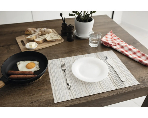 Masă de mic dejun luminoasă cu suport aspect lemn, farfurie albă, tacâmuri, tigaie cu ou și cârnați, pâine și șervețel în carouri.