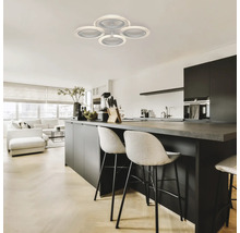 Fotografie de interior a unui spațiu de locuit cu o lampă de tavan cu patru abajururi rotunde, o insulă de bucătărie cu scaune de bar și o sufragerie cu mobilier alb.