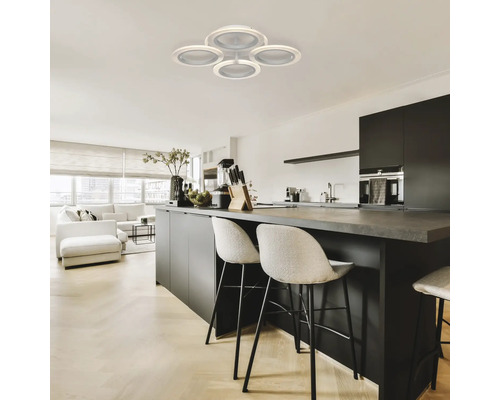 Fotografie de interior a unui spațiu de locuit cu o lampă de tavan cu patru abajururi rotunde, o insulă de bucătărie cu scaune de bar și o sufragerie cu mobilier alb.