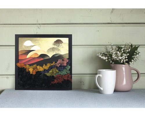 Tablou pătrat în ramă neagră cu peisaj stilizat din mușchi colorat, decorat lângă o cană albă și o vază roz pe o masă.