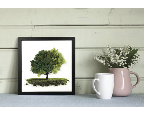 Tablou pătrat din mușchi în ramă neagră cu motiv de copac, lângă o cană albă și o vază roz cu lăcrămioare.