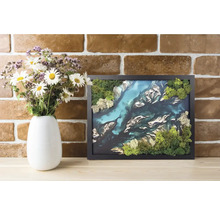 Tablou 3D cu mușchi în ramă neagră cu peisaj de râu din mușchi conservat lângă o vază cu flori în fața unui perete de cărămidă.