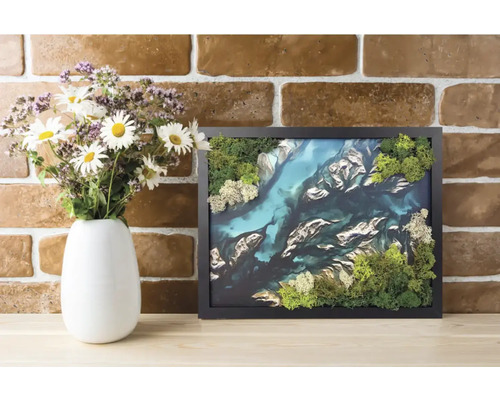 Tablou 3D cu mușchi în ramă neagră cu peisaj de râu din mușchi conservat lângă o vază cu flori în fața unui perete de cărămidă.