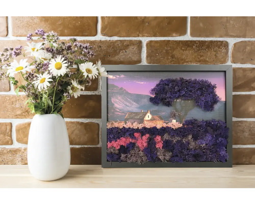 Tablou din mușchi stabilizat cu peisaj, casă și copac în ramă neagră, pe o poliță de lemn lângă vază cu flori în fața unui perete de cărămidă.