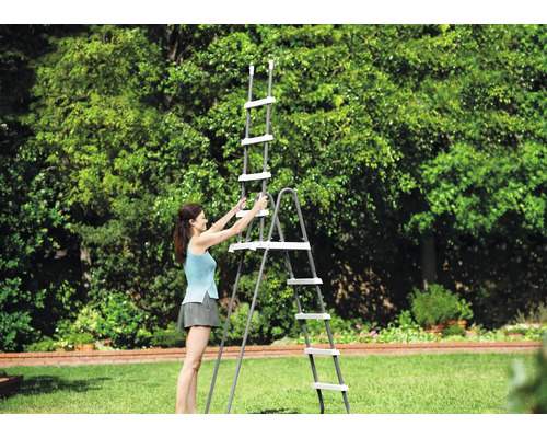 O femeie montează o scară de piscină gri cu trepte albe pe un gazon într-o grădină luminoasă și spațioasă.