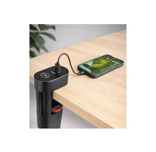 Stație de încărcare USB neagră cu clemă de masă fixată de marginea unui birou, încărcând un smartphone prin cablu pe o suprafață din lemn deschis.