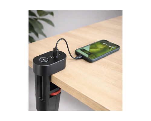Stație de încărcare USB neagră cu clemă de masă fixată de marginea unui birou, încărcând un smartphone prin cablu pe o suprafață din lemn deschis.