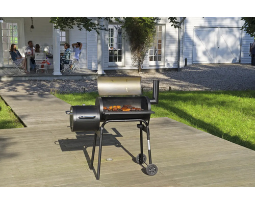 Grătar smoker negru cu capac deschis și carne pe grătar, amplasat pe o terasă din lemn în fața unei case albe.