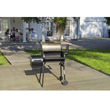 Grătar tip afumătoare negru pe o terasă din lemn în grădină, cu o casă albă și un grup de persoane în fundal.