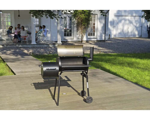 Grătar tip afumătoare negru pe o terasă din lemn în grădină, cu o casă albă și un grup de persoane în fundal.