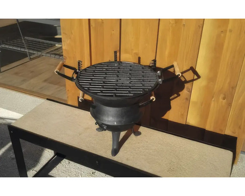 Grătar pe cărbuni din fontă cu trei picioare și mânere din lemn în fața unui perete din lemn.