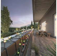 Ghirlandă luminoasă LED colorată pentru exterior, înfășurată pe o balustradă de balcon pe o terasă de lemn cu masă și bănci la amurg.