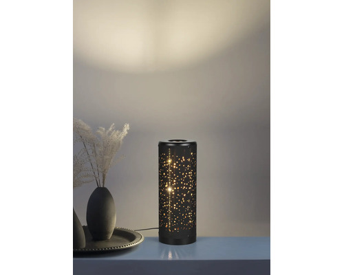 Veioză cilindrică din metal negru cu model de stele perforate și efect de lumină caldă într-un decor.