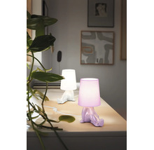 Două veioze decorative în formă de figurină care stă pe o masă deschisă la culoare, una albă și una mov, lângă o plantă verde.