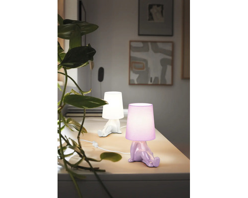 Două veioze decorative în formă de figurină care stă pe o masă deschisă la culoare, una albă și una mov, lângă o plantă verde.