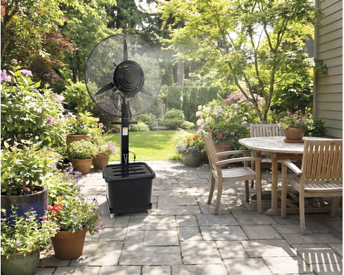 Ventilator cu pulverizare negru pe o terasă pavată cu mobilier de grădină din lemn și numeroase plante cu flori la ghiveci într-o grădină luminoasă.