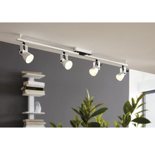 Sistem modern de iluminat pe șină alb cu patru spoturi reglabile pe un tavan într-o cameră luminoasă cu raft pentru cărți și plante.