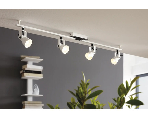 Sistem modern de iluminat pe șină alb cu patru spoturi reglabile pe un tavan într-o cameră luminoasă cu raft pentru cărți și plante.