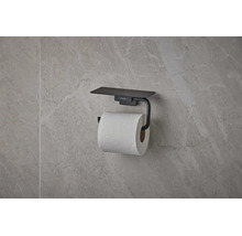 Suport pentru hârtie igienică hansgrohe negru mat cu poliță, montat pe un perete cu aspect de piatră.