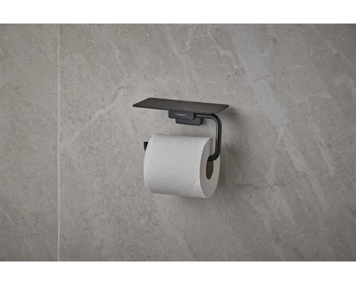 Suport pentru hârtie igienică hansgrohe negru mat cu poliță, montat pe un perete cu aspect de piatră.