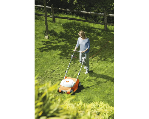 Femeie care utilizează un scarificator electric pe un gazon într-o grădină luminoasă cu gard de lemn.