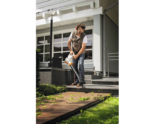Un bărbat cu echipament de protecție curăță o terasă din lemn deschis cu un suflător de frunze electric STIHL în fața unei case moderne.