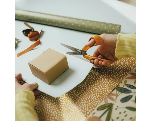 O persoană folosește foarfeca universală Fiskars pentru a tăia hârtie albă pentru ambalarea unei cutii mici de carton.