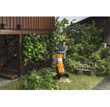 Un bărbat folosește un tocător de grădină Stihl pentru a toca crengi într-o grădină lângă o clădire din lemn.