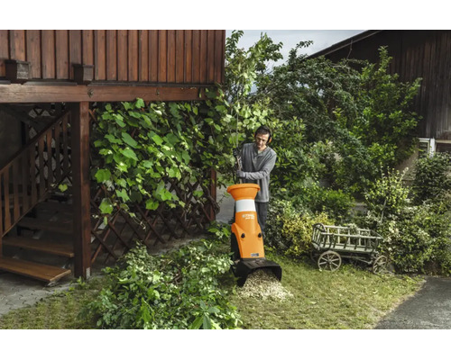 Un bărbat folosește un tocător de grădină Stihl pentru a toca crengi într-o grădină lângă o clădire din lemn.