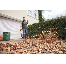 Un bărbat folosește o suflantă de frunze STIHL pentru a curăța frunzele uscate de pe o alee pavată în fața unei case cu garaj și gard viu.