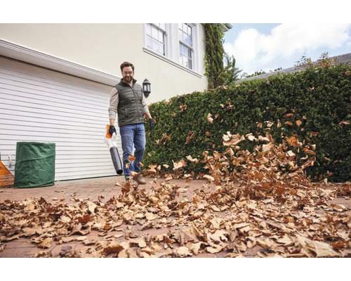 Un bărbat folosește o suflantă de frunze STIHL pentru a curăța frunzele uscate de pe o alee pavată în fața unei case cu garaj și gard viu.
