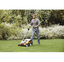 Un bărbat folosește un scarificator Stihl pe un gazon într-o grădină cu tufișuri.