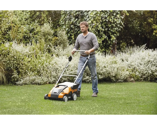Un bărbat folosește un scarificator Stihl pe un gazon într-o grădină cu tufișuri.