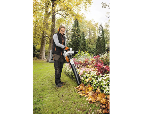 Un bărbat lucrează cu un aspirator de frunze STIHL într-o grădină luminoasă. El aspiră frunzele de toamnă dintr-un strat de flori plantat.