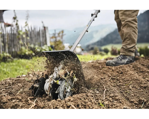 O persoană folosește un accesoriu de motocultor pentru a afâna pământul într-o grădină cu gard și munți în fundal.