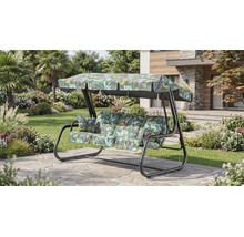 Balansoar de grădină cu 3 locuri cu model tropical de frunze pe perne și acoperiș, pe o terasă din piatră într-o grădină luminoasă și amenajată.