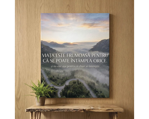 Tablou pe pânză cu peisaj montan, răsărit și citat în română, decorațiune de perete deasupra unei mese cu plantă.