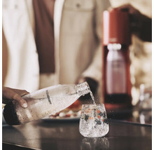 O persoană toarnă apă acidulată dintr-o sticlă SodaStream într-un pahar cu gheață și fructe de pădure. Un aparat de făcut sifon se vede în fundal.