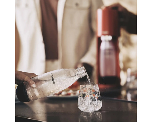 O persoană toarnă apă acidulată dintr-o sticlă SodaStream într-un pahar cu gheață și fructe de pădure. Un aparat de făcut sifon se vede în fundal.