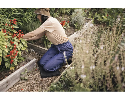 O femeie îngenuncheată pe o pernă pentru genunchi lucrând într-o grădină ridicată din lemn cu diverse plante și flori roșii.