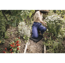 O persoană îngenunchează pe o protecție pentru genunchi în timp ce grădinărește într-un strat cu diverse plante și arbuști înfloriți.