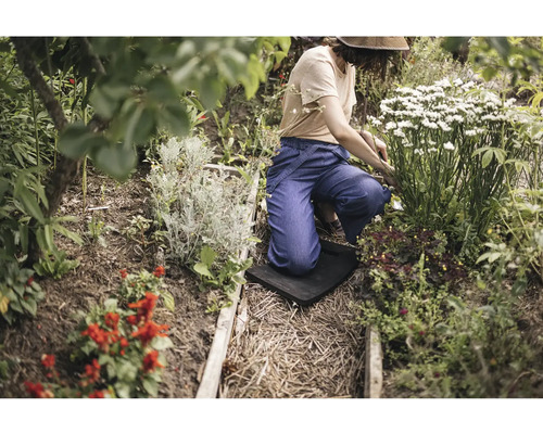 O persoană îngenunchează pe o protecție pentru genunchi în timp ce grădinărește într-un strat cu diverse plante și arbuști înfloriți.