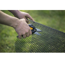 O persoană taie o plasă din plastic negru cu o foarfecă universală Fiskars în grădină.