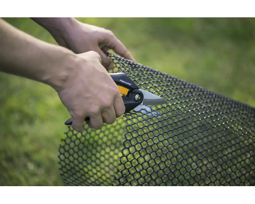 O persoană taie o plasă din plastic negru cu o foarfecă universală Fiskars în grădină.