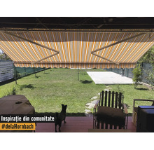Copertină cu braț articulat în dungi galben-albe deasupra unei terase cu vedere spre o grădină verde și mobilier de exterior.