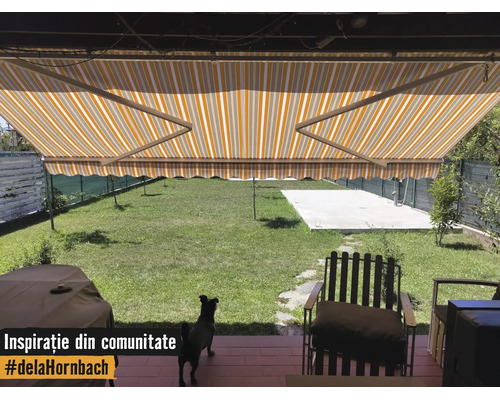 Copertină cu braț articulat în dungi galben-albe deasupra unei terase cu vedere spre o grădină verde și mobilier de exterior.